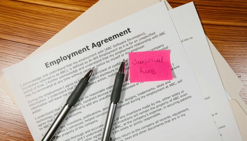 Top view of employment agreement contract with pens and seasonal hire note on wooden desk.