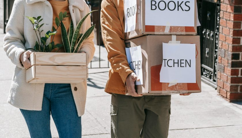 Couple smiling while carrying boxes and plants during urban relocation in daylight.