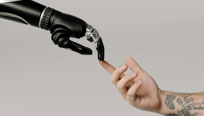 A human hand with tattoos reaching out to a robotic hand on a white background.
